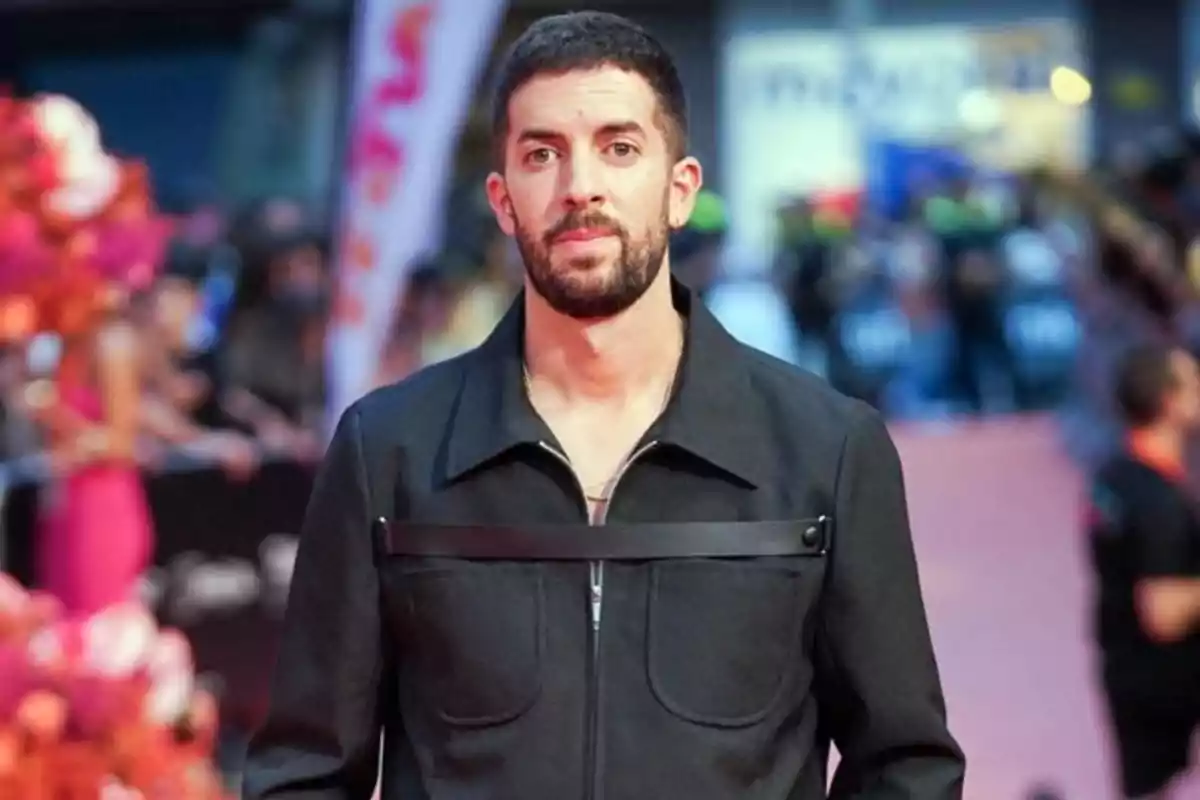 Hombre con barba y cabello corto vestido con chaqueta oscura en una alfombra roja con fondo desenfocado de personas y decoraciones coloridas Hombre con barba y cabello corto vestido con chaqueta oscura en una alfombra roja con fondo desenfocado de personas y decoraciones coloridas