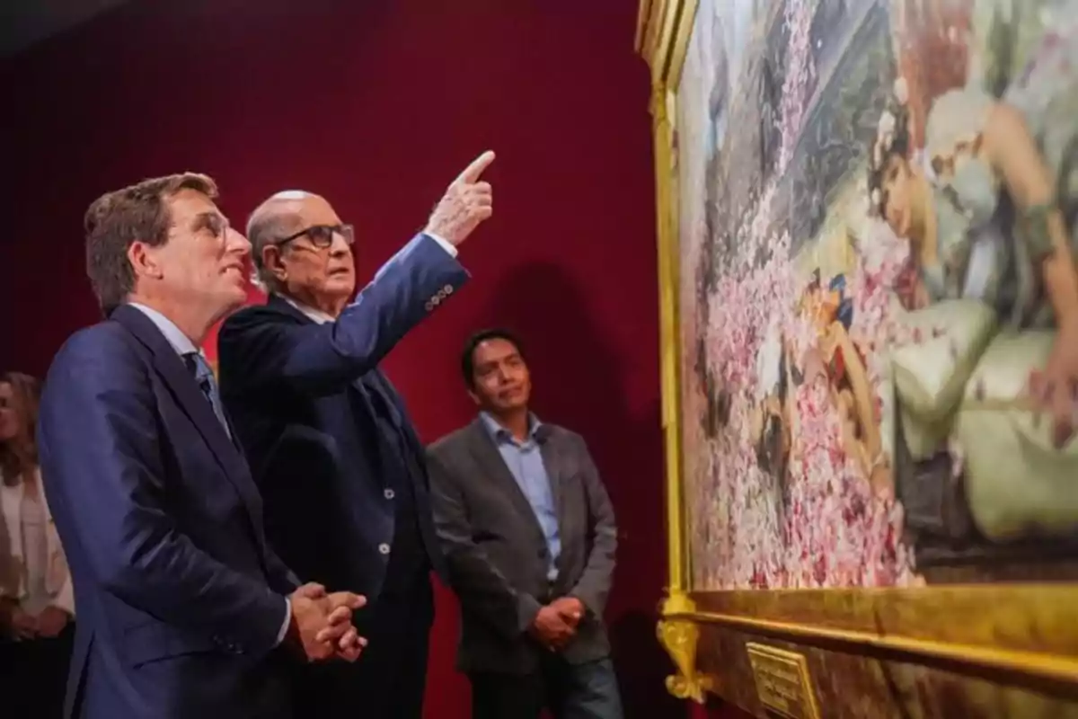 Dos hombres de traje observan atentamente una pintura enmarcada en un museo mientras uno de ellos señala un detalle del cuadro y otras personas los acompañan al fondo Dos hombres de traje observan atentamente una pintura enmarcada en un museo mientras uno de ellos señala un detalle del cuadro y otras personas los acompañan al fondo