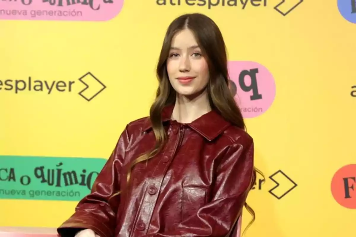 Joven con cabello largo y ondulado lleva una chaqueta de cuero roja y posa frente a un fondo amarillo con logotipos y texto de colores Joven con cabello largo y ondulado lleva una chaqueta de cuero roja y posa frente a un fondo amarillo con logotipos y texto de colores