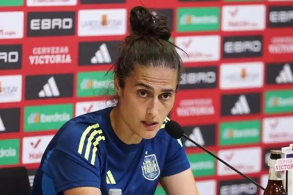 Entrenadora de fútbol con camiseta azul de la selección española hablando en una rueda de prensa frente a un micrófono con fondo de patrocinadores Entrenadora de fútbol con camiseta azul de la selección española hablando en una rueda de prensa frente a un micrófono con fondo de patrocinadores
