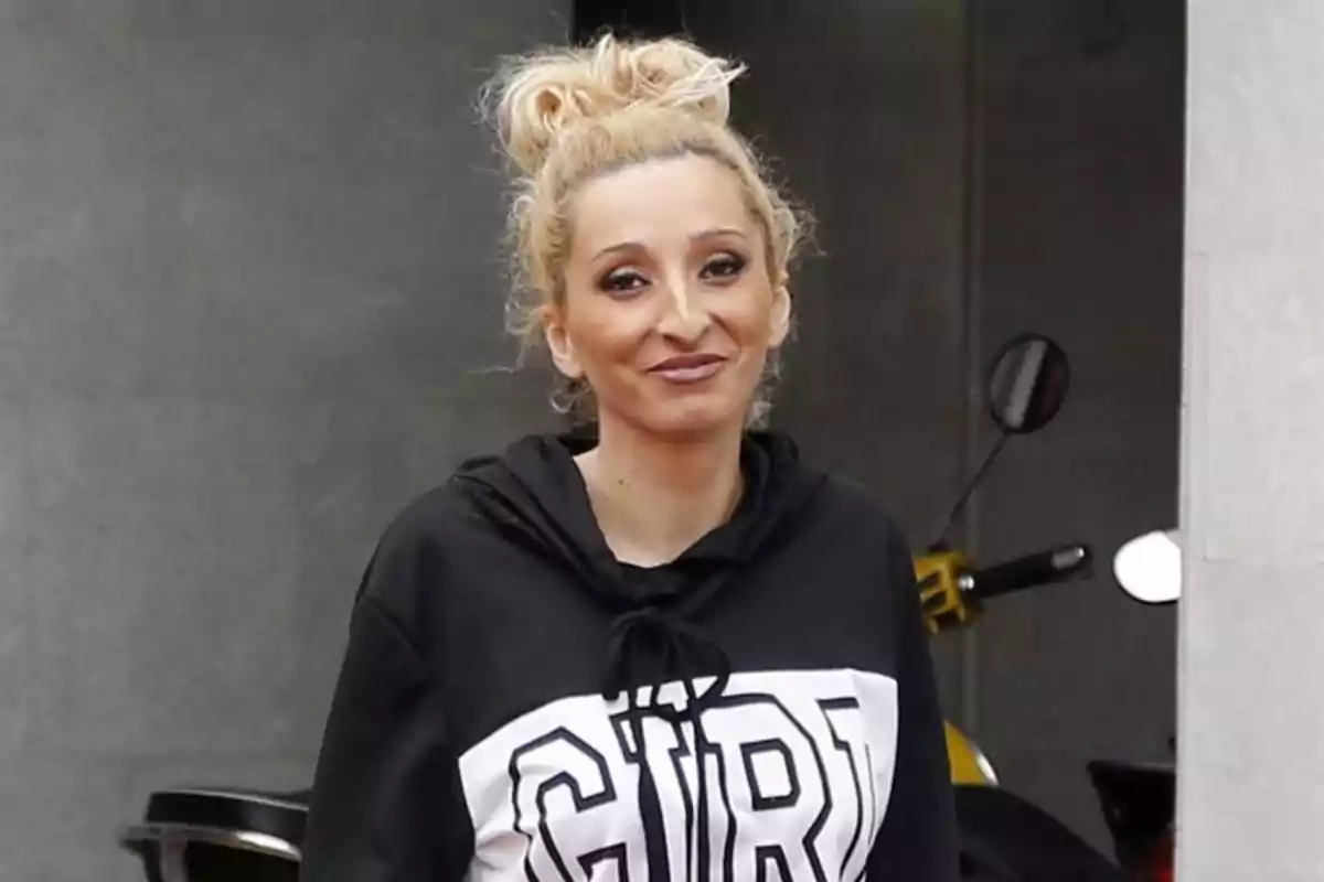 Mujer rubia con el cabello recogido en un moño y sudadera negra con letras blancas sonríe frente a una pared gris Mujer rubia con el cabello recogido en un moño y sudadera negra con letras blancas sonríe frente a una pared gris