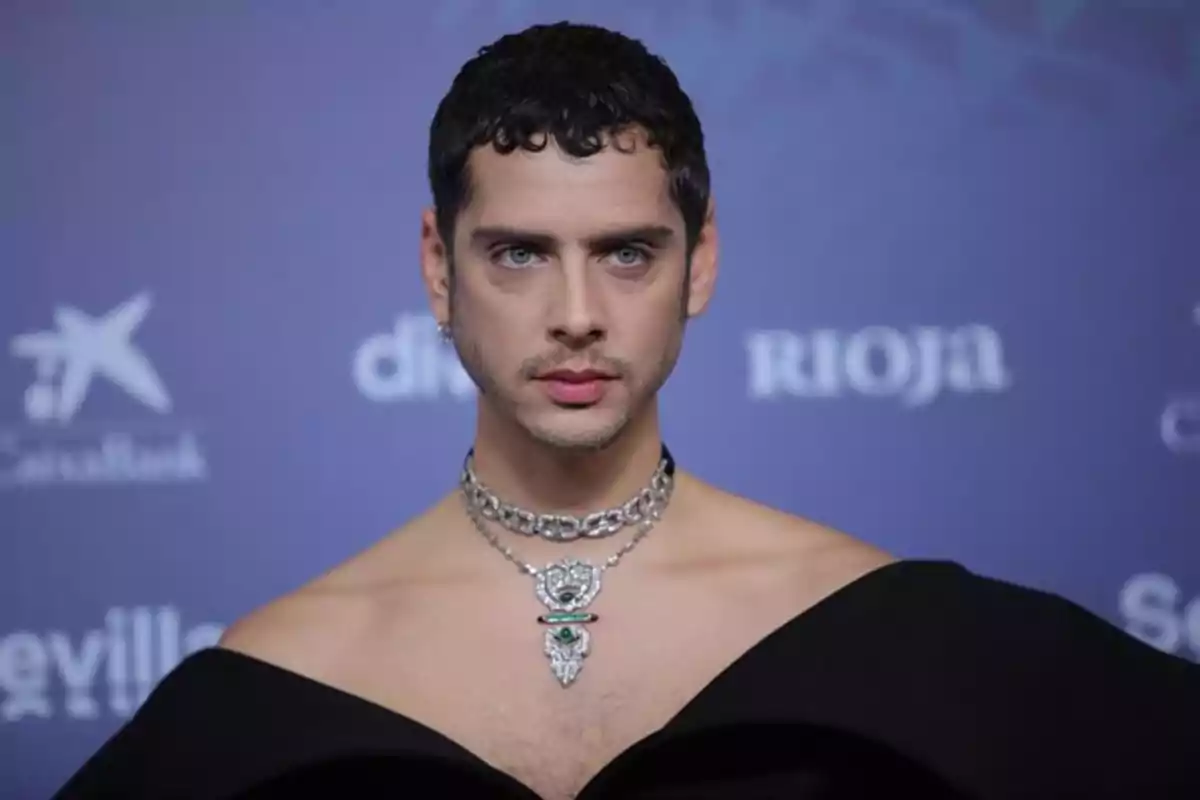 Persona de cabello corto y oscuro con maquillaje y labios pintados usando un collar llamativo y un atuendo elegante de hombros descubiertos posando frente a un fondo azul con logotipos desenfocados.