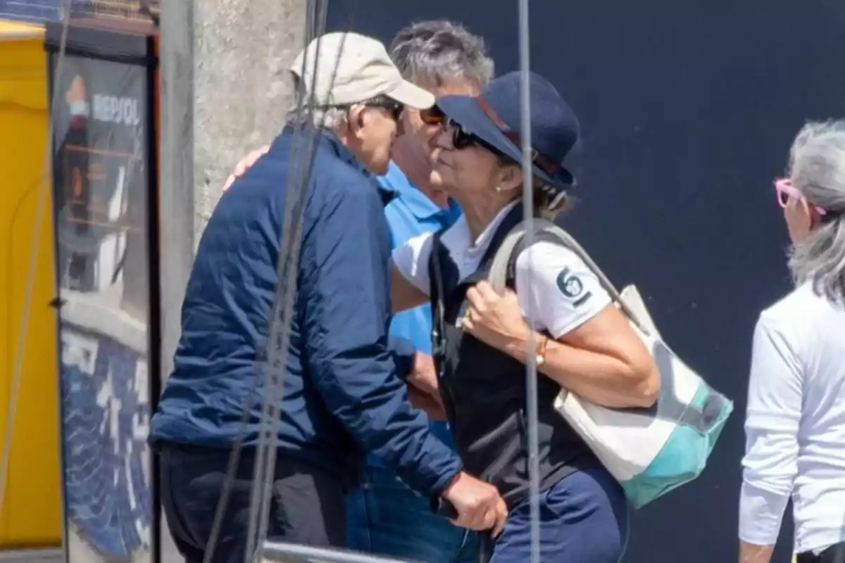 Two people greet each other with a kiss on the cheek in an outdoor setting, both wearing casual clothes and sunglasses, while other people are nearby. Two people greet each other with a kiss on the cheek in an outdoor setting, both wearing casual clothes and sunglasses, while other people are nearby.