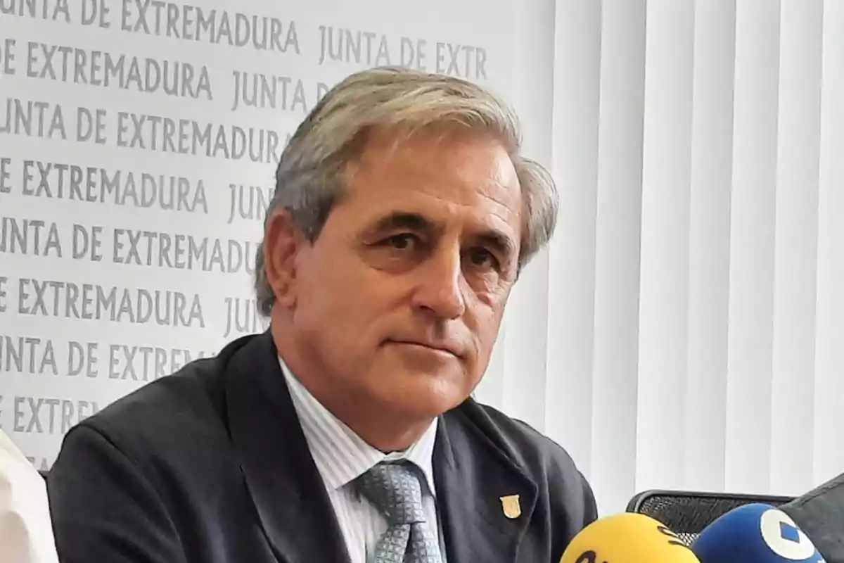 Hombre de cabello canoso con traje y corbata sentado frente a micrófonos en una rueda de prensa con fondo de la Junta de Extremadura Hombre de cabello canoso con traje y corbata sentado frente a micrófonos en una rueda de prensa con fondo de la Junta de Extremadura
