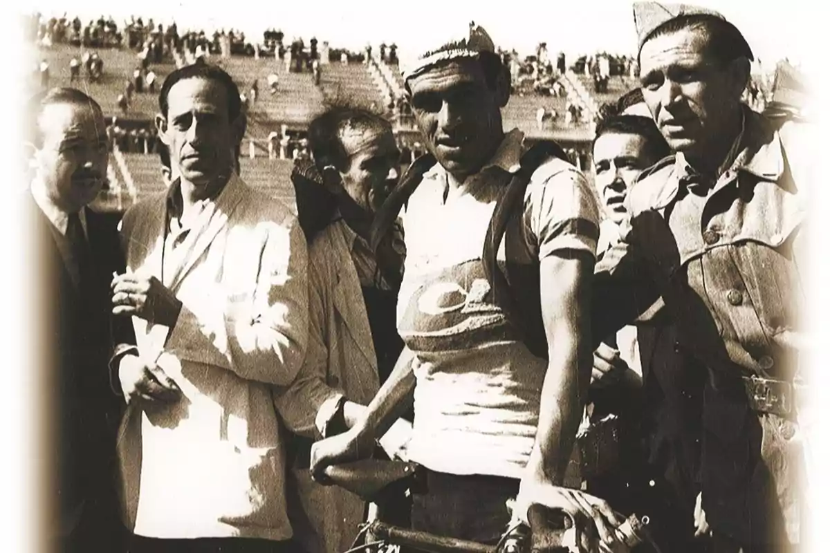 Grupo de hombres rodeando a un ciclista con bicicleta en un estadio antiguo lleno de espectadores al fondo Grupo de hombres rodeando a un ciclista con bicicleta en un estadio antiguo lleno de espectadores al fondo
