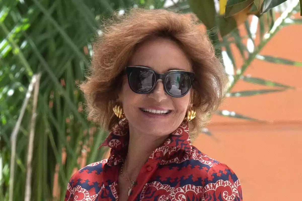 Mujer sonriente con gafas de sol, blusa estampada en rojo y azul y pendientes dorados, posando al aire libre frente a plantas verdes Mujer sonriente con gafas de sol, blusa estampada en rojo y azul y pendientes dorados, posando al aire libre frente a plantas verdes