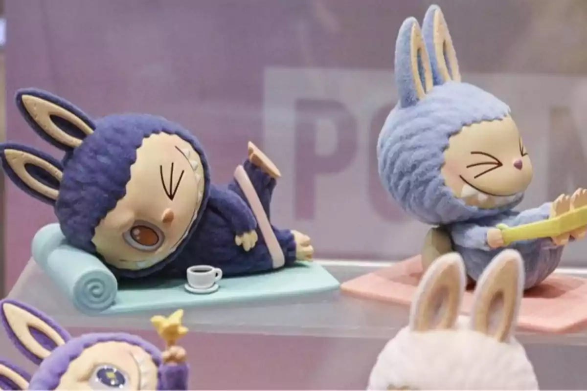Dos figuras de juguete con orejas de conejo y trajes de peluche, una recostada sobre una colchoneta con una taza y la otra sentada sonriendo y estirando las piernas
