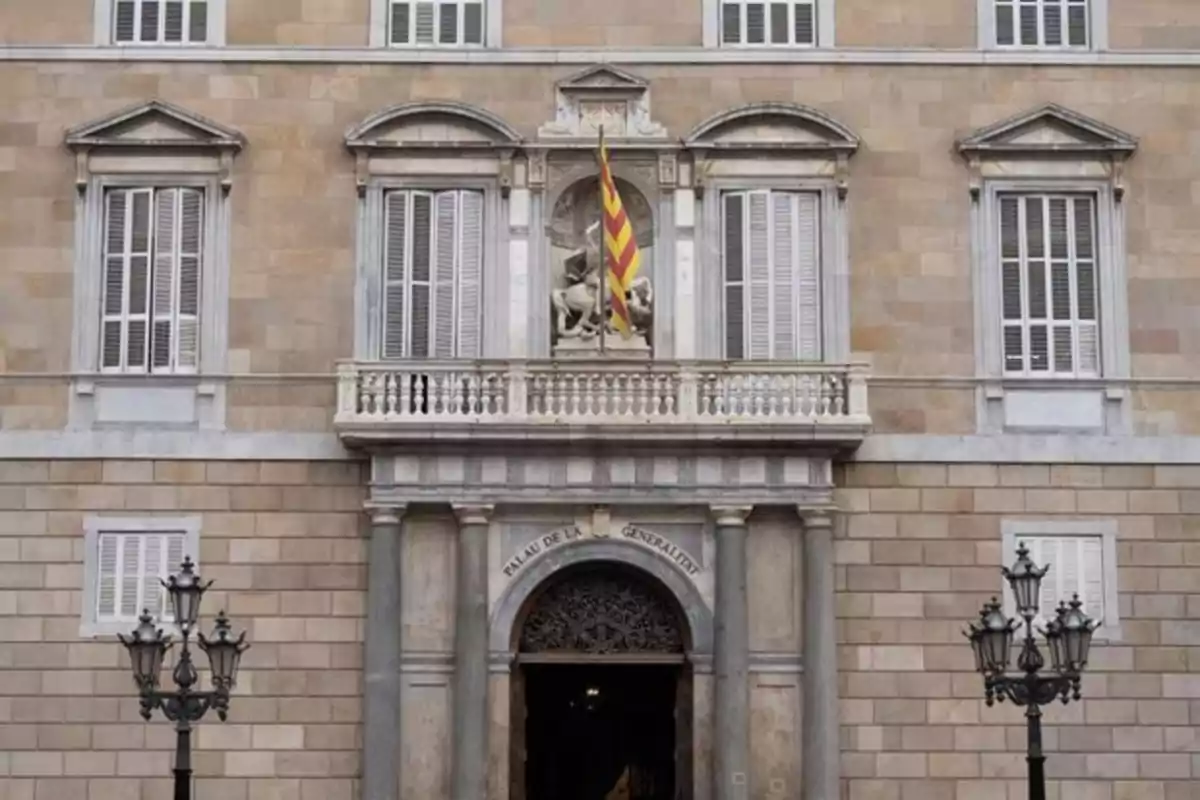 Fachada principal de un edificio histórico con ventanas blancas, balcón central, escultura y bandera catalana ondeando Fachada principal de un edificio histórico con ventanas blancas, balcón central, escultura y bandera catalana ondeando