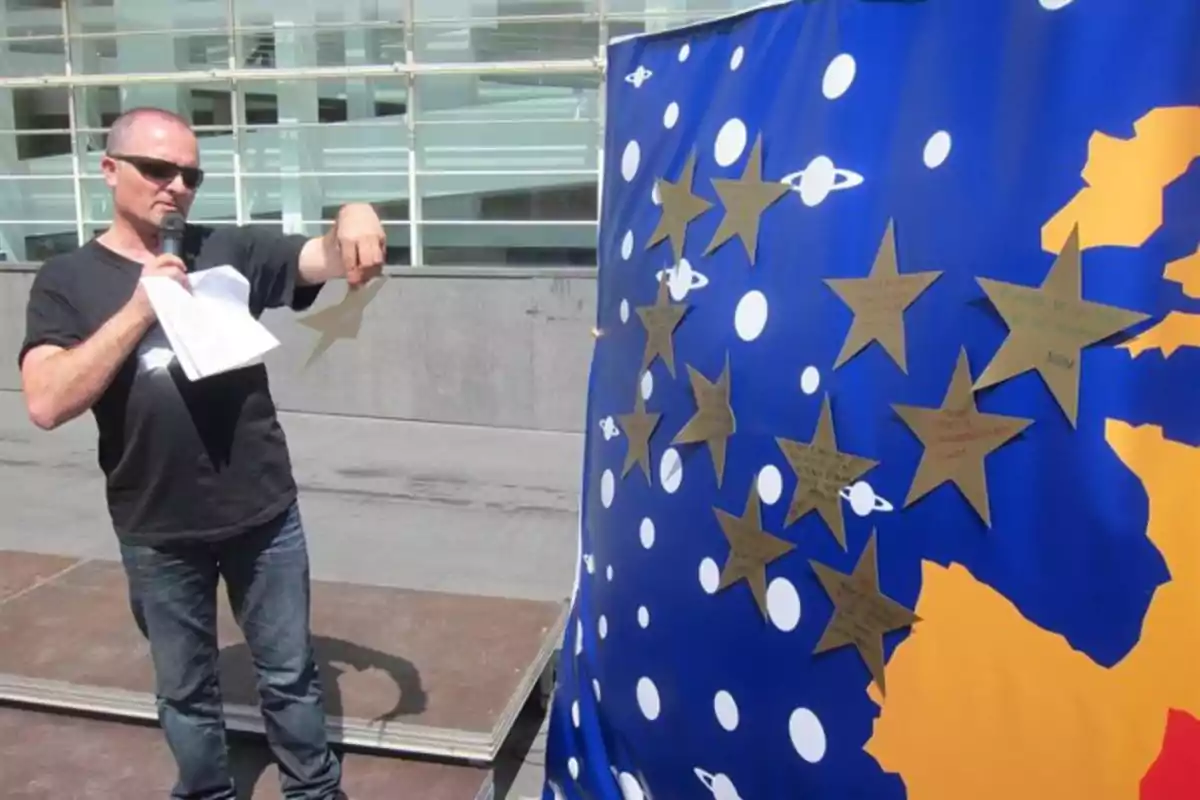 Hombre con gafas de sol sosteniendo un micrófono y una estrella de papel frente a un mural azul decorado con estrellas doradas y planetas blancos Hombre con gafas de sol sosteniendo un micrófono y una estrella de papel frente a un mural azul decorado con estrellas doradas y planetas blancos