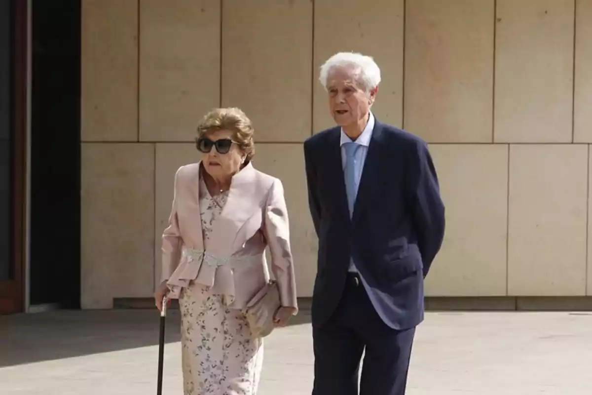 Una pareja de personas mayores elegantemente vestidas camina junta al aire libre frente a una pared de piedra clara Una pareja de personas mayores elegantemente vestidas camina junta al aire libre frente a una pared de piedra clara