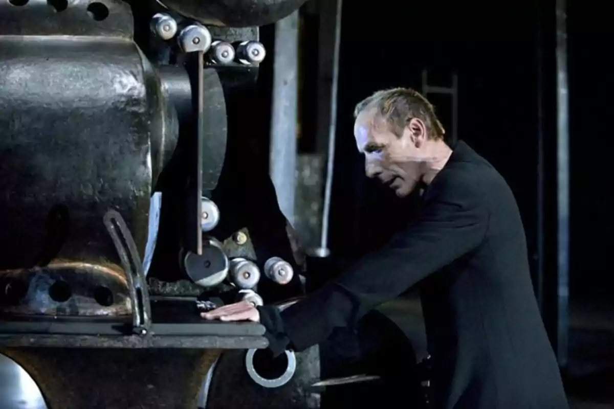 Hombre de perfil vestido de negro apoyando las manos sobre una gran máquina industrial en un ambiente oscuro Hombre de perfil vestido de negro apoyando las manos sobre una gran máquina industrial en un ambiente oscuro