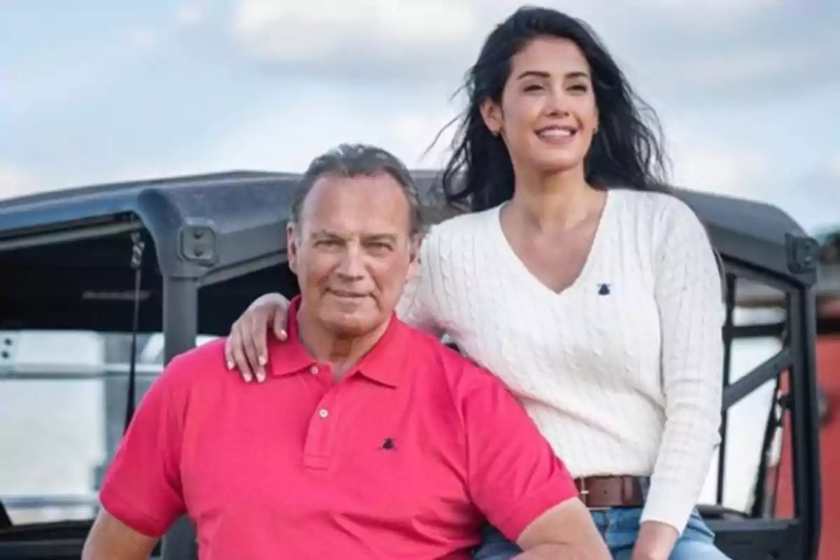 Hombre de cabello canoso con camiseta polo rosa y mujer de cabello largo y oscuro con suéter blanco posan juntos al aire libre frente a un vehículo negro