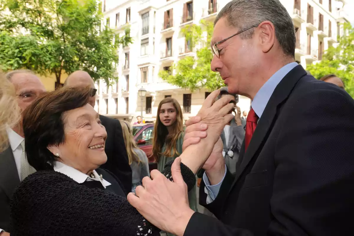 Un hombre con gafas y traje saluda afectuosamente a una mujer mayor que sonríe mientras varias personas observan la escena en una calle con árboles y edificios al fondo Un hombre con gafas y traje saluda afectuosamente a una mujer mayor que sonríe mientras varias personas observan la escena en una calle con árboles y edificios al fondo