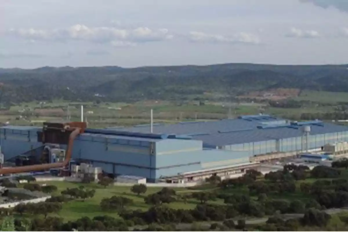 Vista panorámica de una gran fábrica industrial azul rodeada de áreas verdes y montañas al fondo Vista panorámica de una gran fábrica industrial azul rodeada de áreas verdes y montañas al fondo
