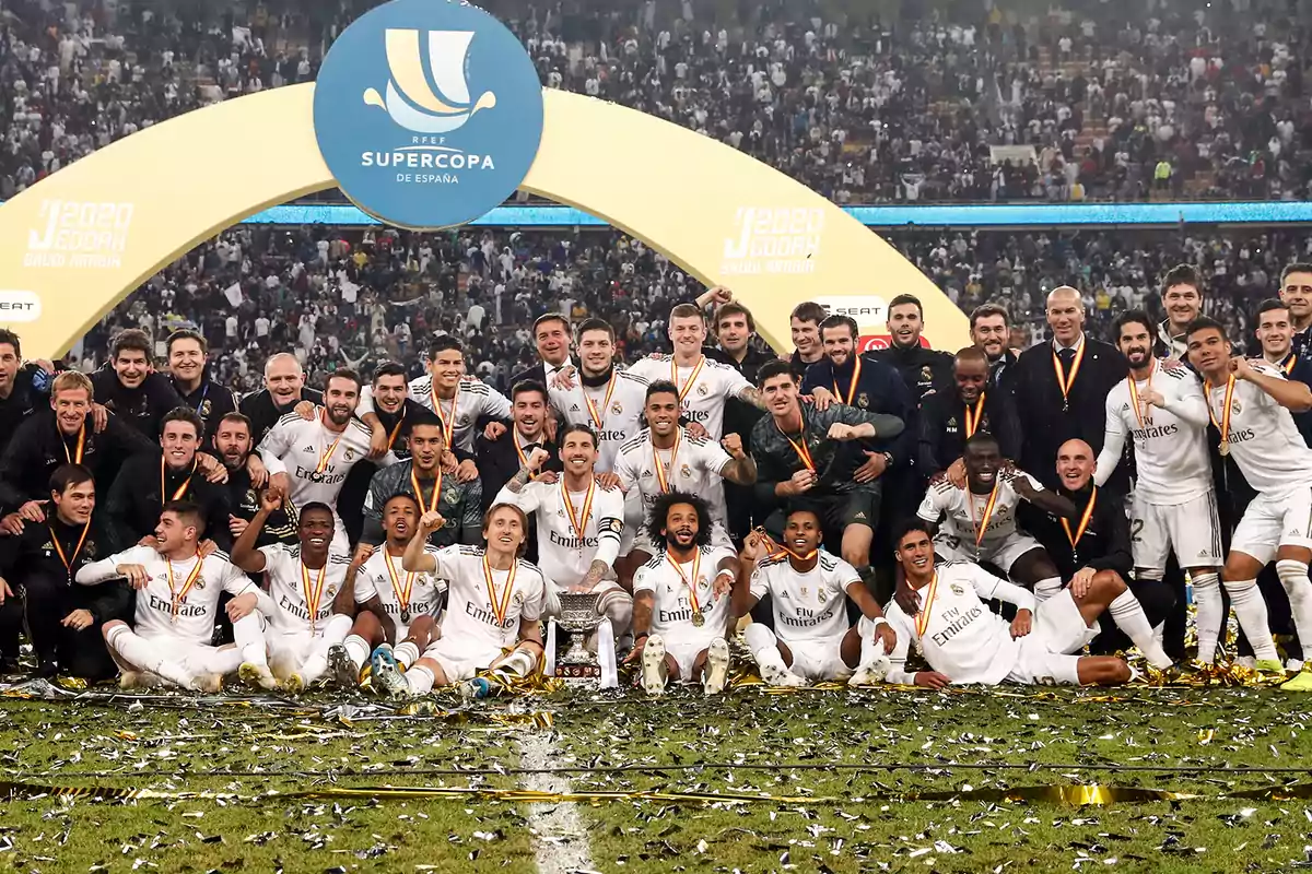Jugadores y cuerpo técnico de un equipo de fútbol celebran juntos en el campo tras ganar la Supercopa de España rodeados de confeti y medallas frente a una multitud en el estadio Jugadores y cuerpo técnico de un equipo de fútbol celebran juntos en el campo tras ganar la Supercopa de España rodeados de confeti y medallas frente a una multitud en el estadio