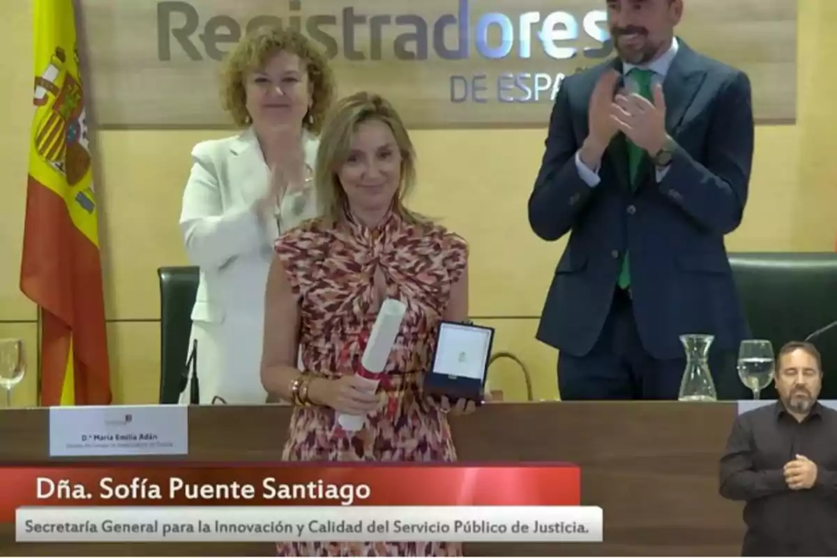 Una mujer sostiene un diploma y una medalla mientras dos personas aplauden detrás de ella en un acto oficial con una bandera de España al fondo y un intérprete de lengua de signos en la esquina inferior derecha