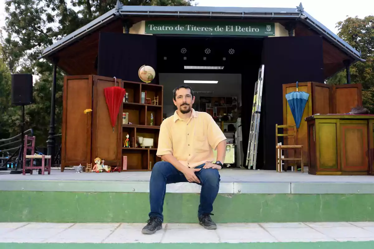 Un hombre con camisa clara y pantalón de mezclilla está sentado en el borde de un escenario al aire libre con decoraciones de teatro y marionetas, al fondo se ve un letrero que dice Teatro de Títeres de El Retiro Un hombre con camisa clara y pantalón de mezclilla está sentado en el borde de un escenario al aire libre con decoraciones de teatro y marionetas, al fondo se ve un letrero que dice Teatro de Títeres de El Retiro