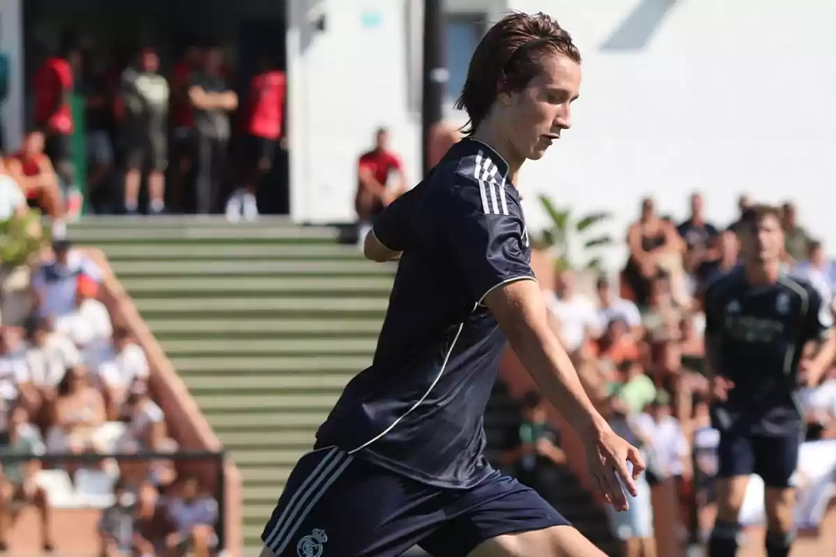 Jugador de fútbol con uniforme oscuro en acción durante un partido, con público y escaleras al fondo Jugador de fútbol con uniforme oscuro en acción durante un partido, con público y escaleras al fondo