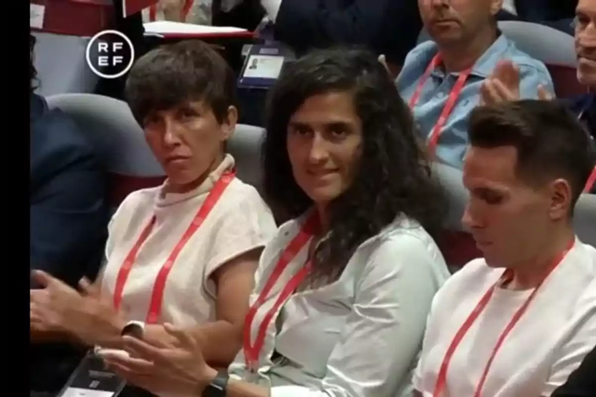 Tres personas sentadas en una conferencia aplaudiendo con expresiones serias y credenciales colgadas al cuello Tres personas sentadas en una conferencia aplaudiendo con expresiones serias y credenciales colgadas al cuello