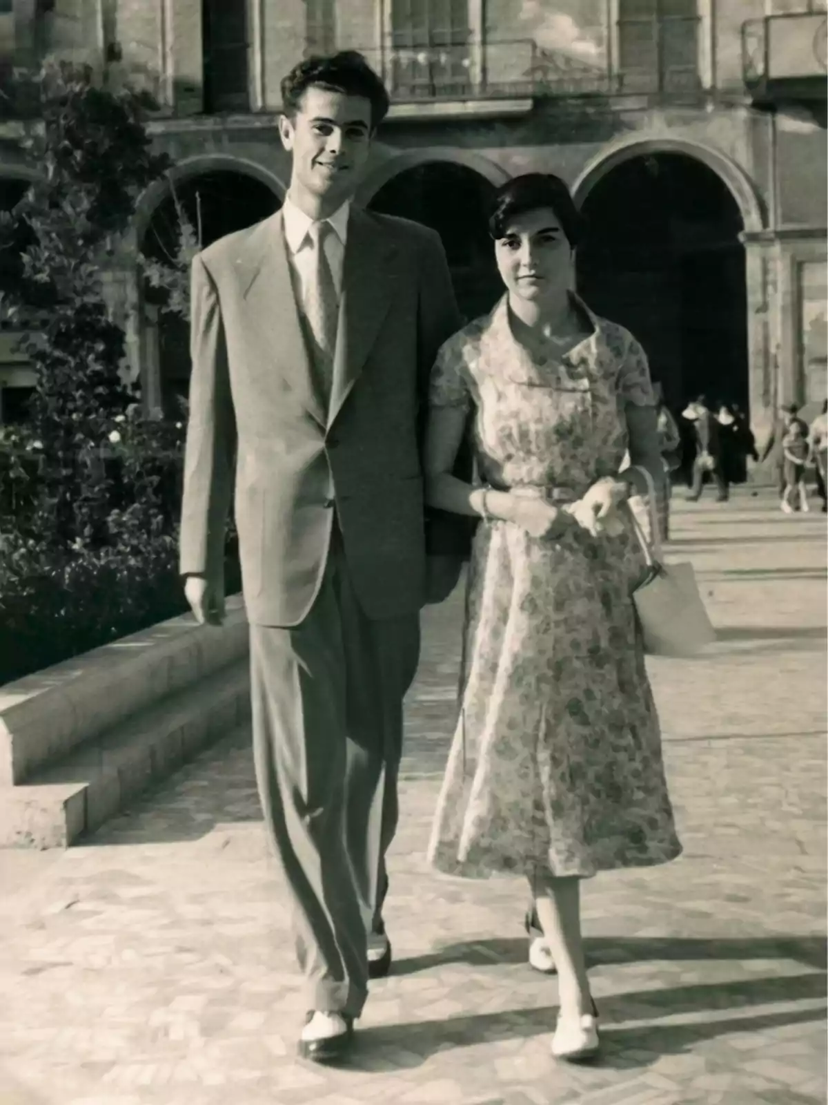 Pareja joven caminando de la mano en una plaza antigua, ambos vestidos de manera elegante y sonrientes Pareja joven caminando de la mano en una plaza antigua, ambos vestidos de manera elegante y sonrientes