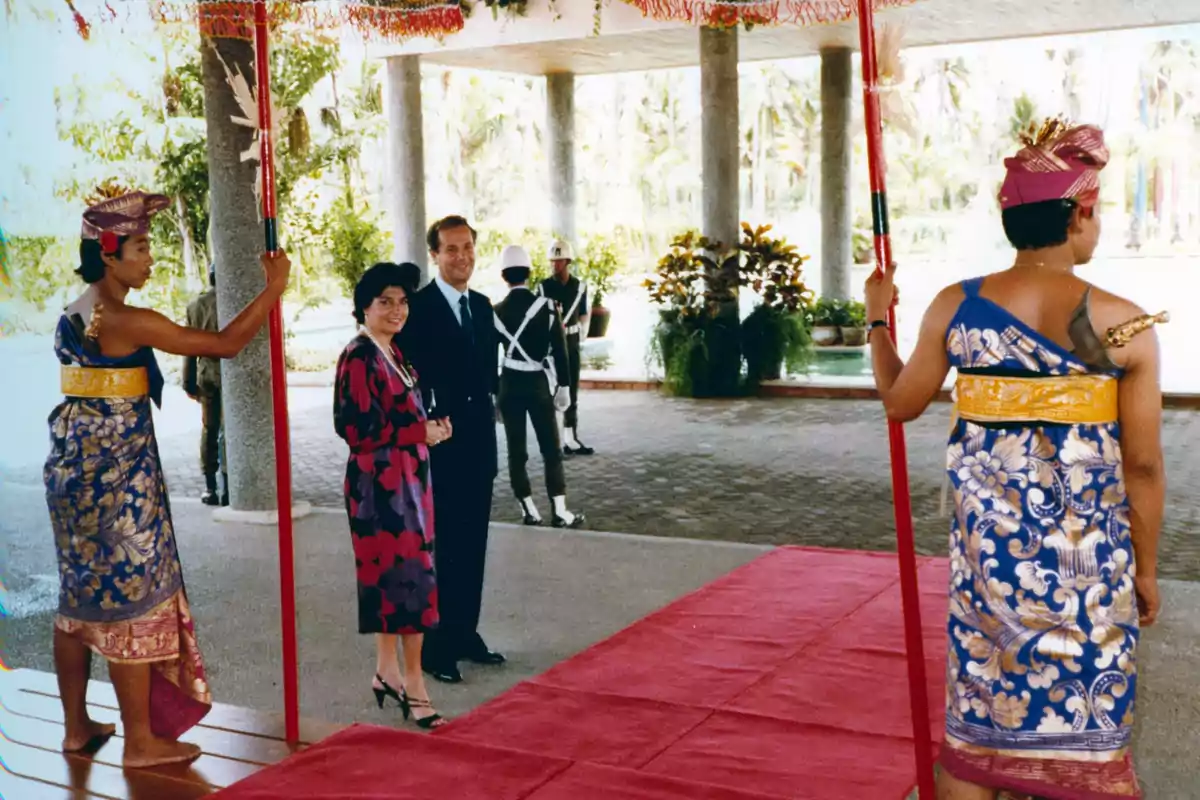 Dos personas elegantemente vestidas caminan sobre una alfombra roja bajo un toldo sostenido por dos hombres con vestimenta tradicional y colorida mientras en el fondo se observan guardias y vegetación Dos personas elegantemente vestidas caminan sobre una alfombra roja bajo un toldo sostenido por dos hombres con vestimenta tradicional y colorida mientras en el fondo se observan guardias y vegetación