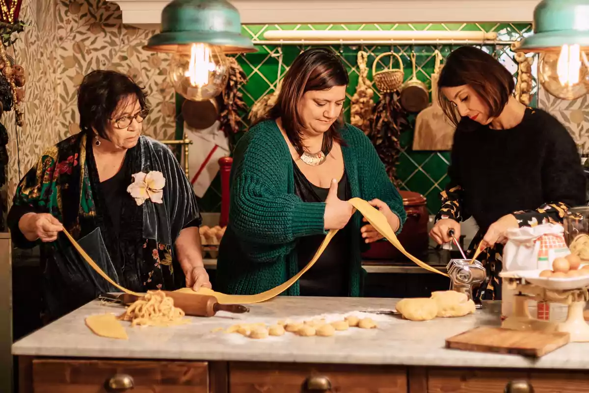 Tres mujeres preparan pasta casera en una cocina acogedora, extendiendo y cortando la masa sobre una mesa de mármol rodeadas de ingredientes y utensilios culinarios.