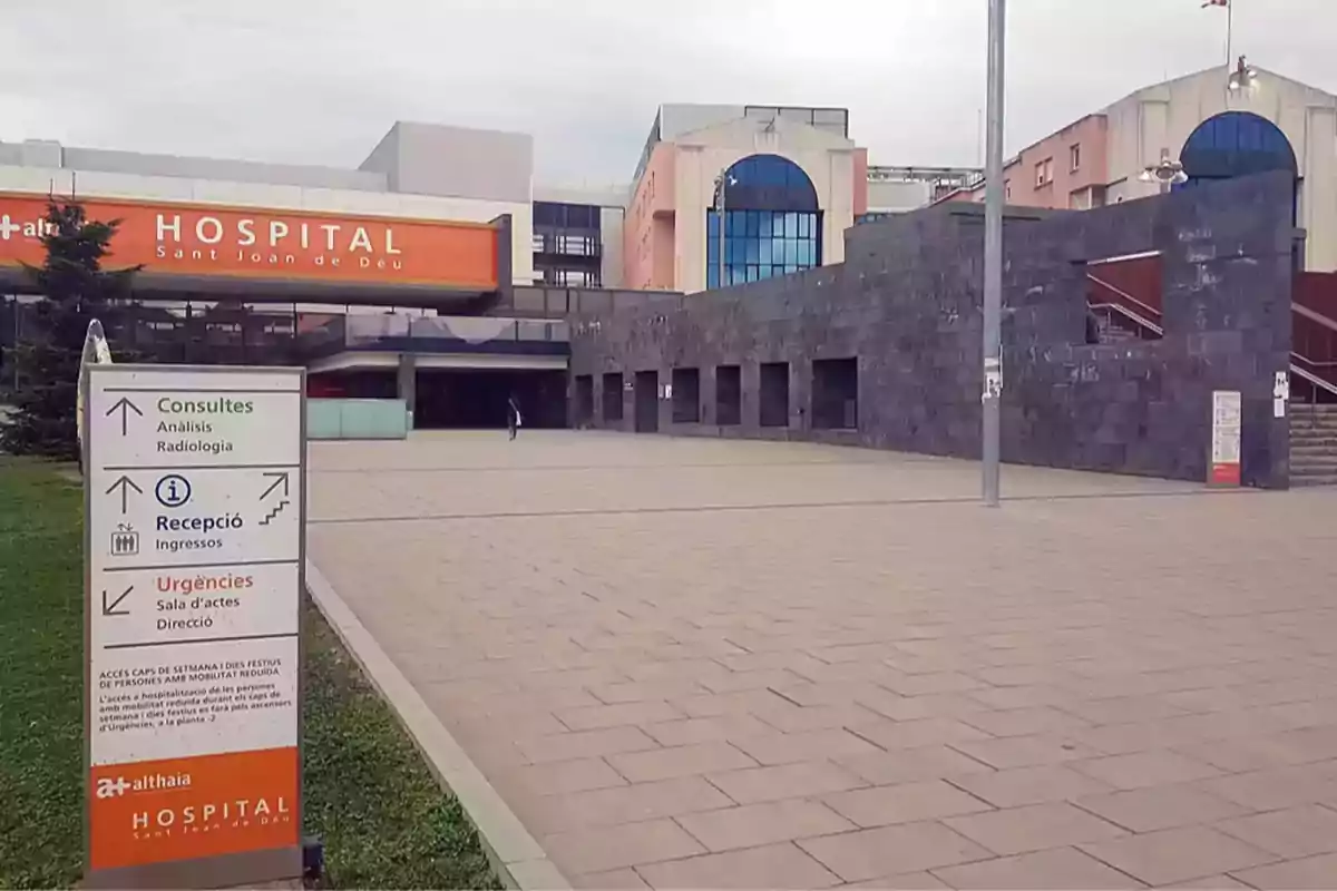 Entrada principal del Hospital Sant Joan de Déu con un cartel de señalización de servicios médicos y urgencias en primer plano Entrada principal del Hospital Sant Joan de Déu con un cartel de señalización de servicios médicos y urgencias en primer plano
