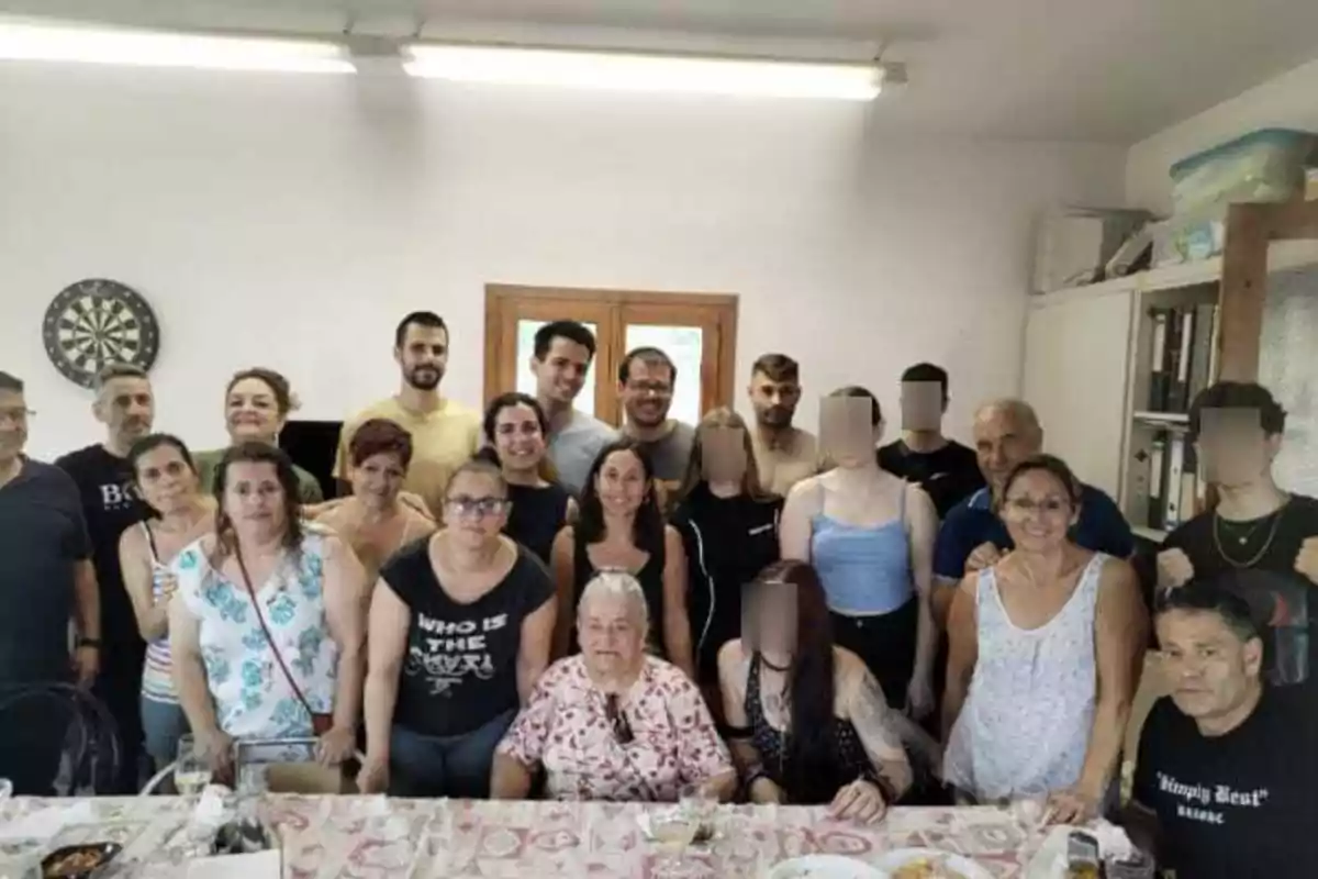 Un grupo grande de personas de diferentes edades posando juntas en una habitación alrededor de una mesa con comida, algunas personas están sentadas y otras de pie, varias tienen el rostro difuminado. Un grupo grande de personas de diferentes edades posando juntas en una habitación alrededor de una mesa con comida, algunas personas están sentadas y otras de pie, varias tienen el rostro difuminado.