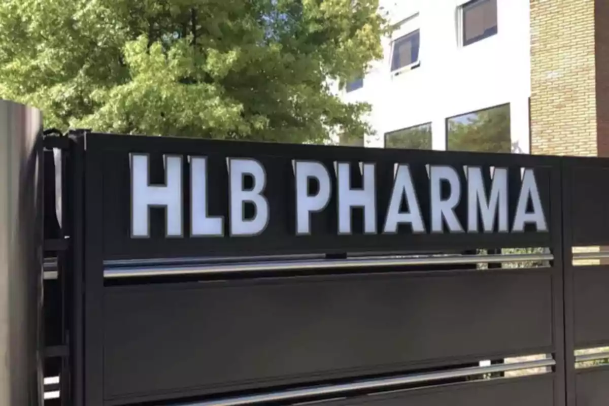 Letrero de HLB Pharma en una reja negra frente a un edificio con ventanas y árboles al fondo Letrero de HLB Pharma en una reja negra frente a un edificio con ventanas y árboles al fondo