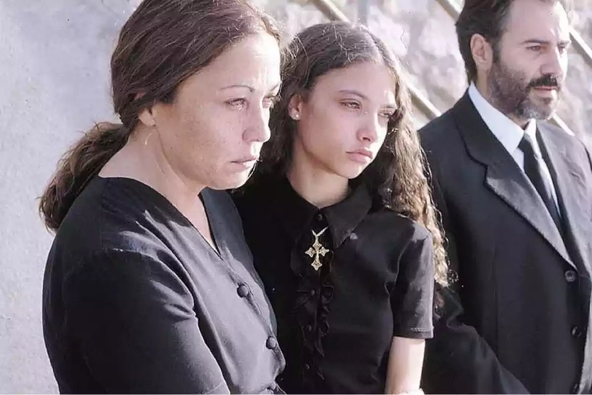 Tres personas vestidas de negro con expresiones serias y tristes, probablemente en un funeral. Tres personas vestidas de negro con expresiones serias y tristes, probablemente en un funeral.