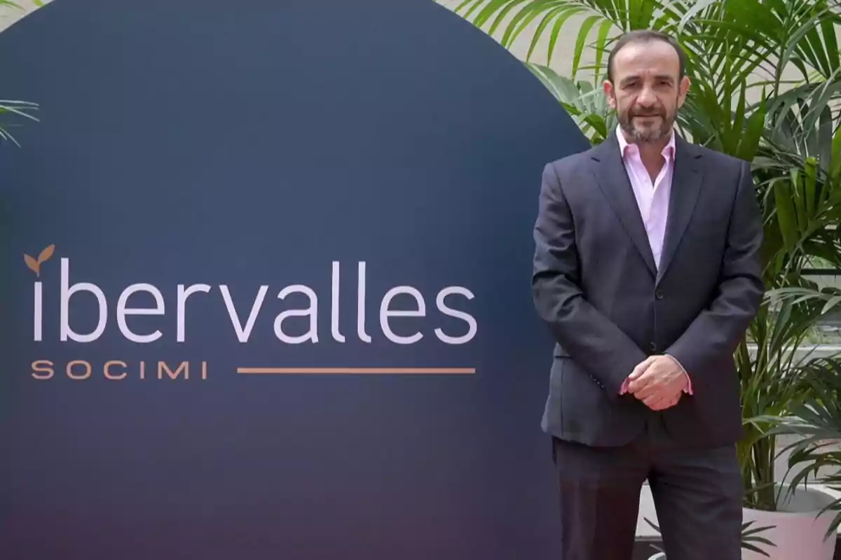 Hombre con barba y traje oscuro posando junto a un cartel de Ibervalles SOCIMI rodeado de plantas verdes Hombre con barba y traje oscuro posando junto a un cartel de Ibervalles SOCIMI rodeado de plantas verdes