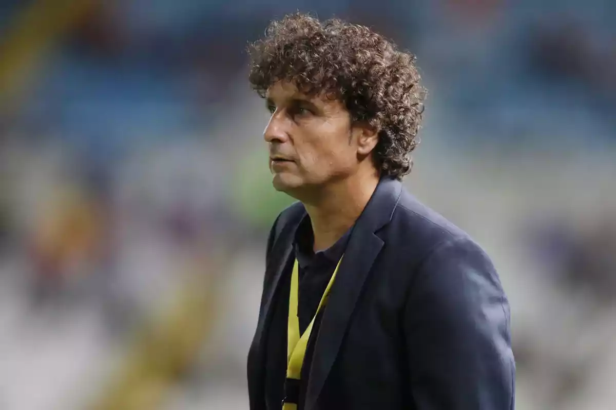 Hombre de cabello rizado con saco azul oscuro y gafete amarillo en un estadio Hombre de cabello rizado con saco azul oscuro y gafete amarillo en un estadio