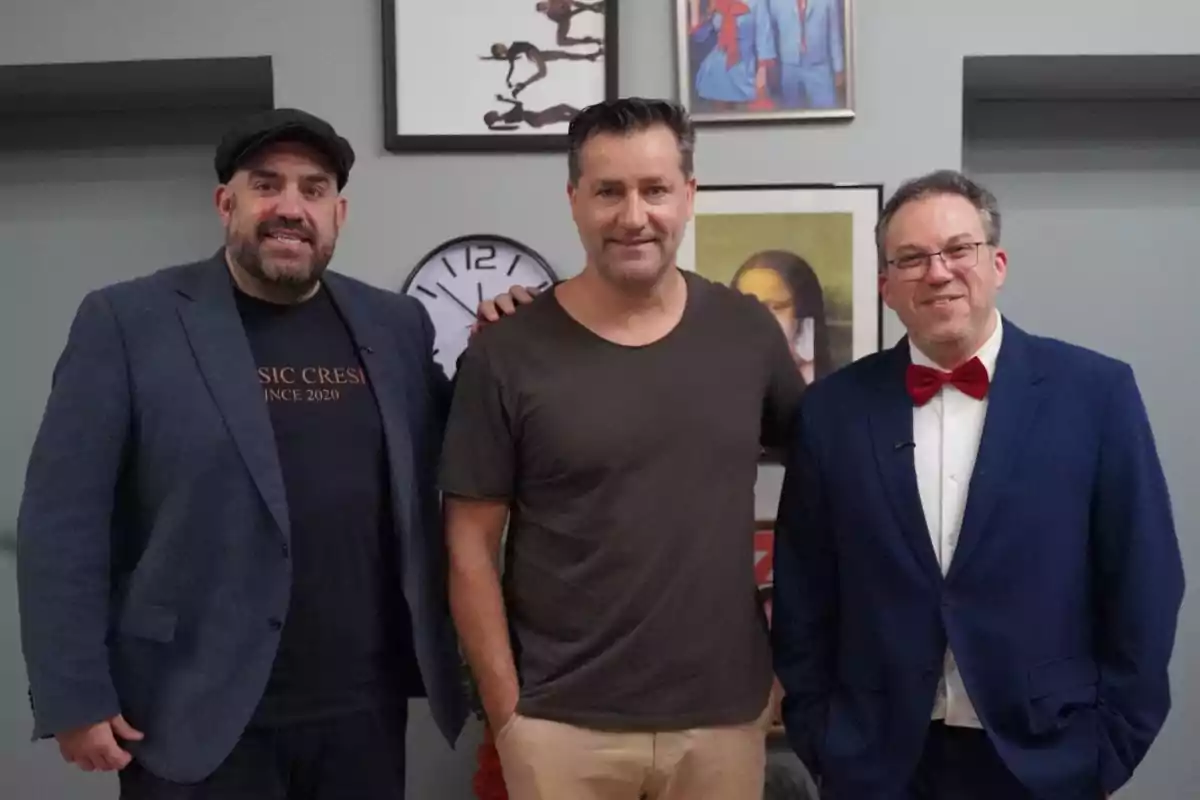 Tres hombres posan juntos en una habitación con paredes grises y varias obras de arte y un reloj en la pared al fondo uno lleva saco azul y gorra otro viste camiseta marrón y pantalón claro y el tercero usa traje azul con moño rojo