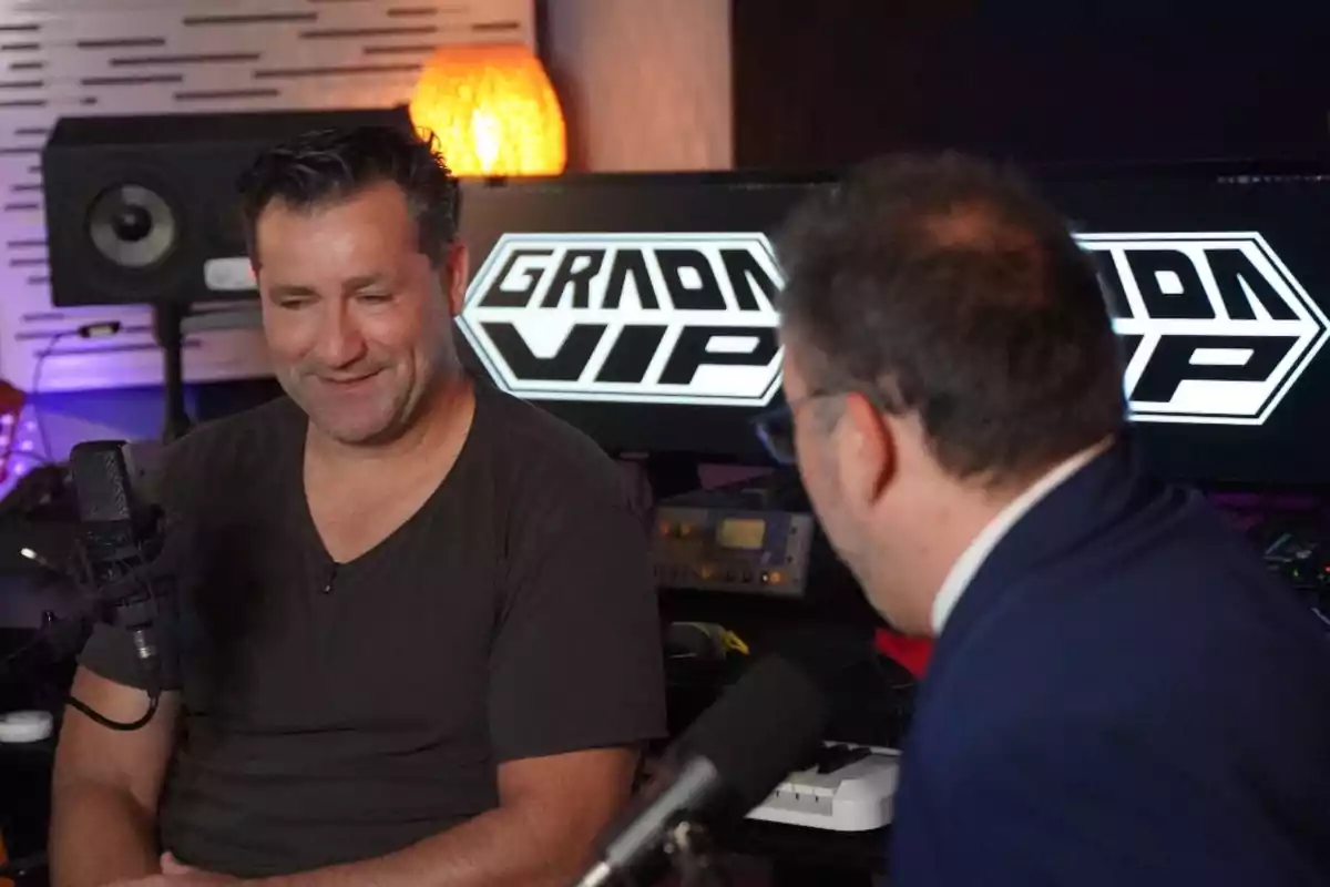 Dos hombres conversan frente a micrófonos en un estudio de grabación con pantallas al fondo que muestran el texto GRADA VIP Dos hombres conversan frente a micrófonos en un estudio de grabación con pantallas al fondo que muestran el texto GRADA VIP