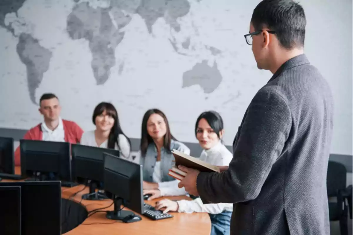 Un grupo de personas sentadas frente a computadoras escucha a un hombre de pie que sostiene libros en un aula con un mapa mundial en la pared.