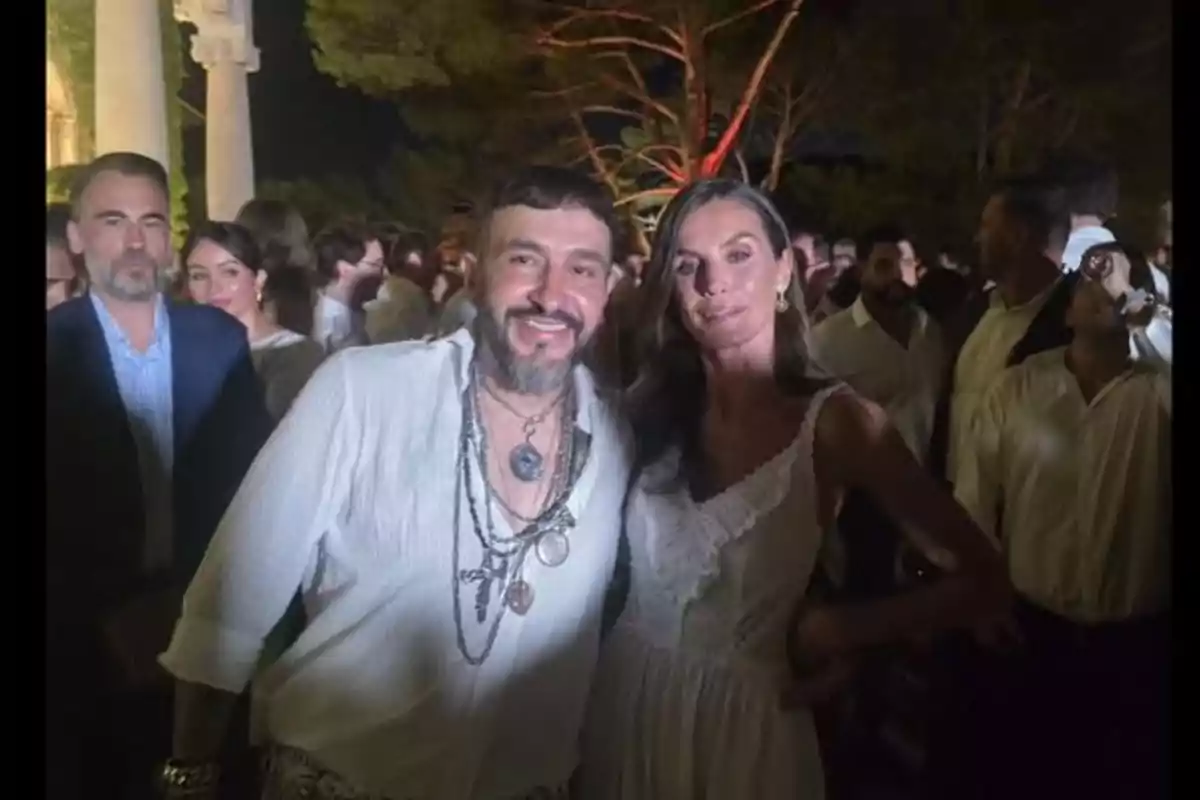 Un hombre y una mujer posan sonrientes en una fiesta nocturna al aire libre rodeados de otras personas, ambos vestidos de blanco y con árboles iluminados al fondo