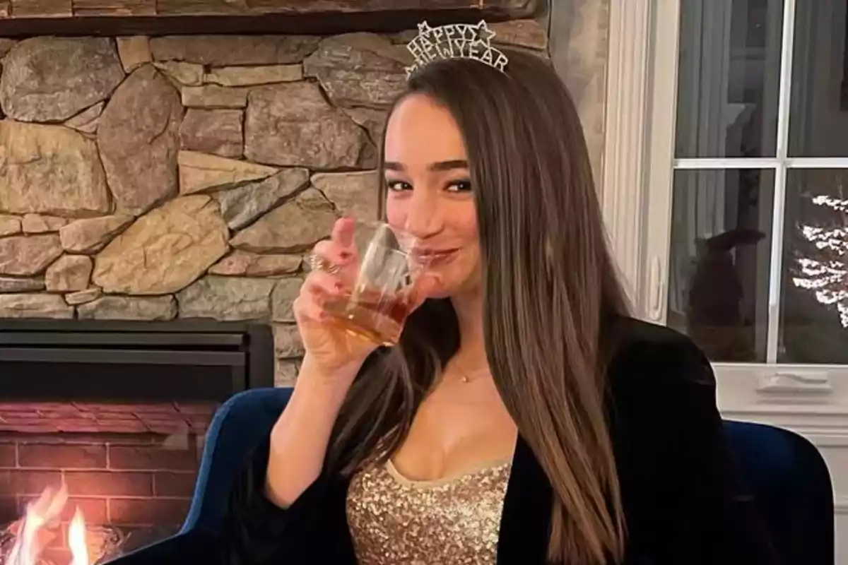 Mujer con diadema de feliz año nuevo sentada junto a una chimenea mientras sostiene un vaso y sonríe Mujer con diadema de feliz año nuevo sentada junto a una chimenea mientras sostiene un vaso y sonríe