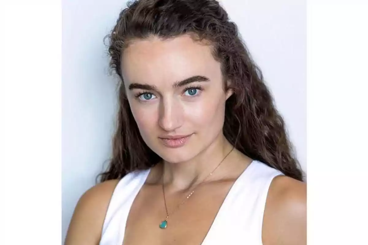 Mujer de cabello rizado y largo con ojos claros, lleva un collar dorado con dije verde y viste una blusa blanca, fondo blanco. Mujer de cabello rizado y largo con ojos claros, lleva un collar dorado con dije verde y viste una blusa blanca, fondo blanco.