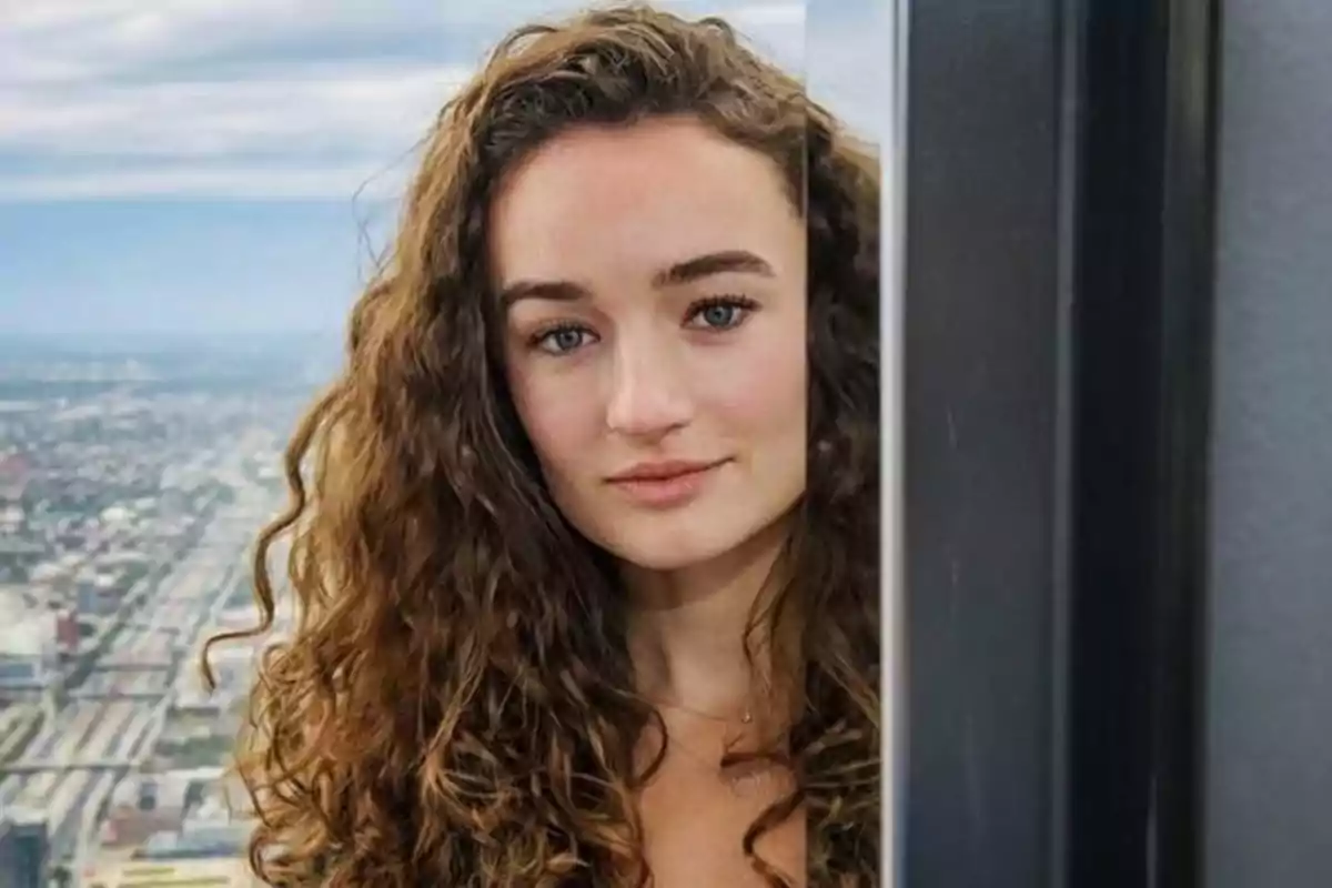 Mujer de cabello rizado y ojos claros mirando a la cámara con una ciudad de fondo y un espejo que refleja la mitad de su rostro