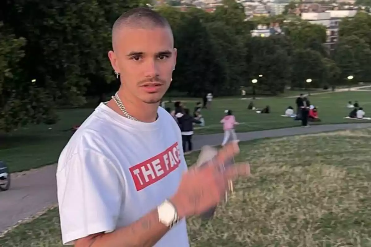 Joven con camiseta blanca y cadena plateada posando en un parque con personas al fondo disfrutando del área verde