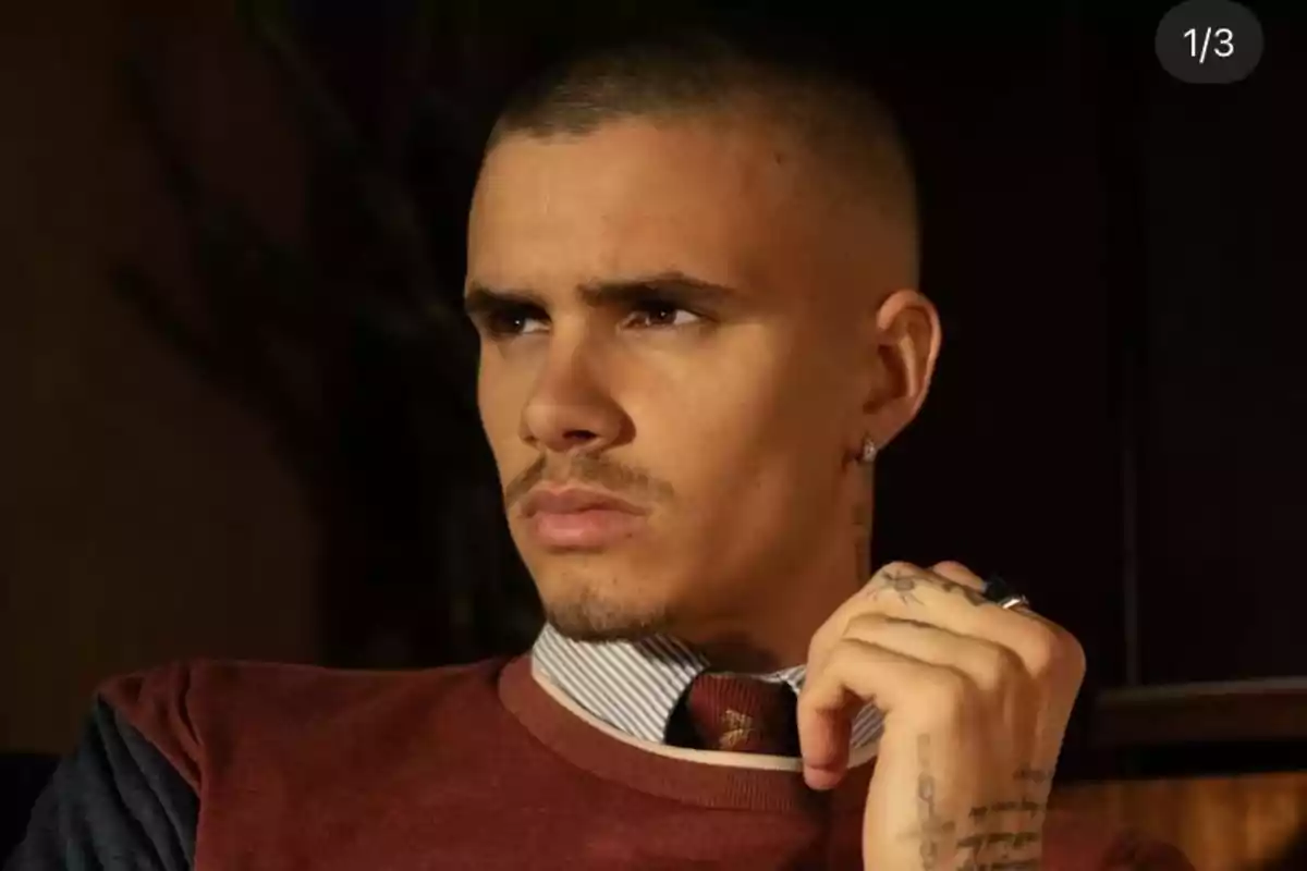Joven con cabello corto y expresión seria mirando hacia un lado lleva un suéter marrón sobre una camisa de rayas y corbata roja tiene tatuajes en la mano y usa un pendiente en la oreja