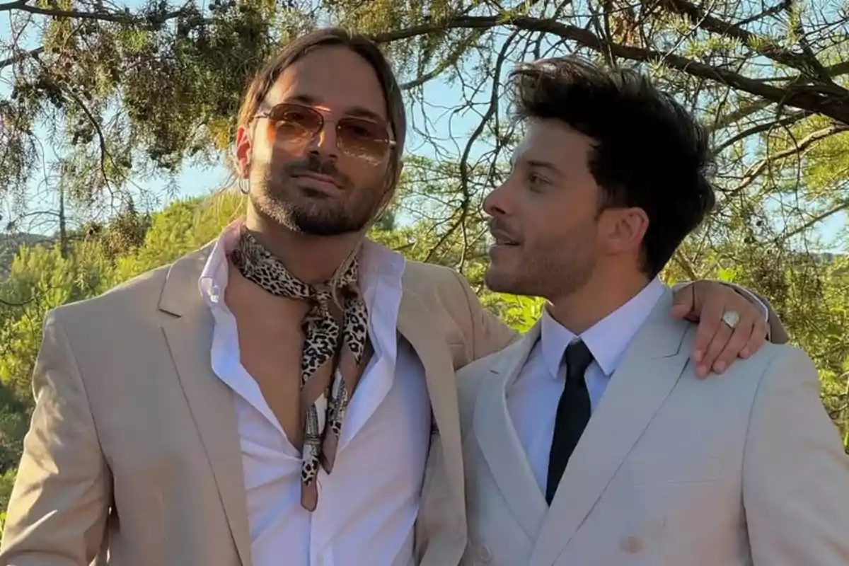Dos hombres vestidos con trajes claros posan juntos al aire libre rodeados de árboles, uno lleva gafas de sol y una bufanda de estampado animal, el otro viste corbata negra y ambos parecen estar disfrutando el momento. Dos hombres vestidos con trajes claros posan juntos al aire libre rodeados de árboles, uno lleva gafas de sol y una bufanda de estampado animal, el otro viste corbata negra y ambos parecen estar disfrutando el momento.
