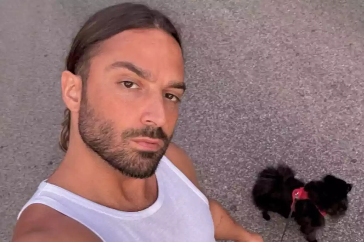 Hombre con barba y camiseta blanca tomándose una selfie mientras pasea a un perro pequeño de pelaje oscuro con arnés rojo sobre el asfalto Hombre con barba y camiseta blanca tomándose una selfie mientras pasea a un perro pequeño de pelaje oscuro con arnés rojo sobre el asfalto