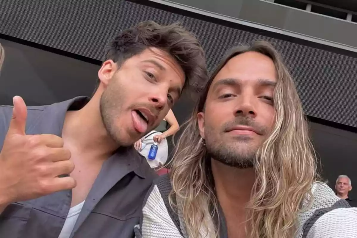 Dos hombres posan para una selfie, uno de ellos sonríe y hace una señal de pulgar arriba mientras el otro tiene el cabello largo y suelto