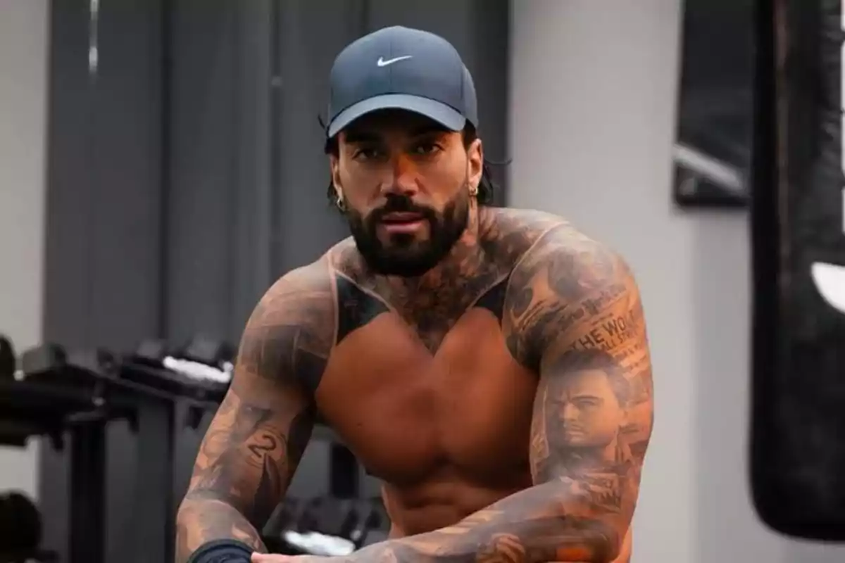 Hombre musculoso con barba y tatuajes en los brazos, usando gorra azul y sentado en un gimnasio Hombre musculoso con barba y tatuajes en los brazos, usando gorra azul y sentado en un gimnasio