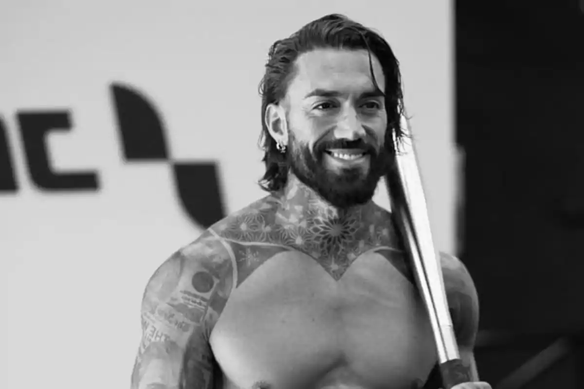 Hombre musculoso con barba y tatuajes sonríe mientras sostiene una barra de metal Hombre musculoso con barba y tatuajes sonríe mientras sostiene una barra de metal
