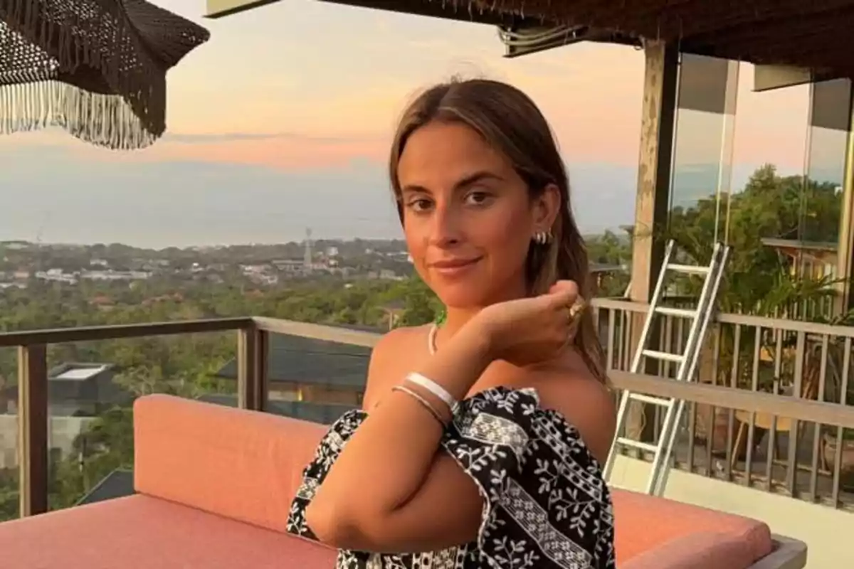 Mujer joven sentada en un sofá al aire libre durante el atardecer con vista a un paisaje de árboles y casas al fondo