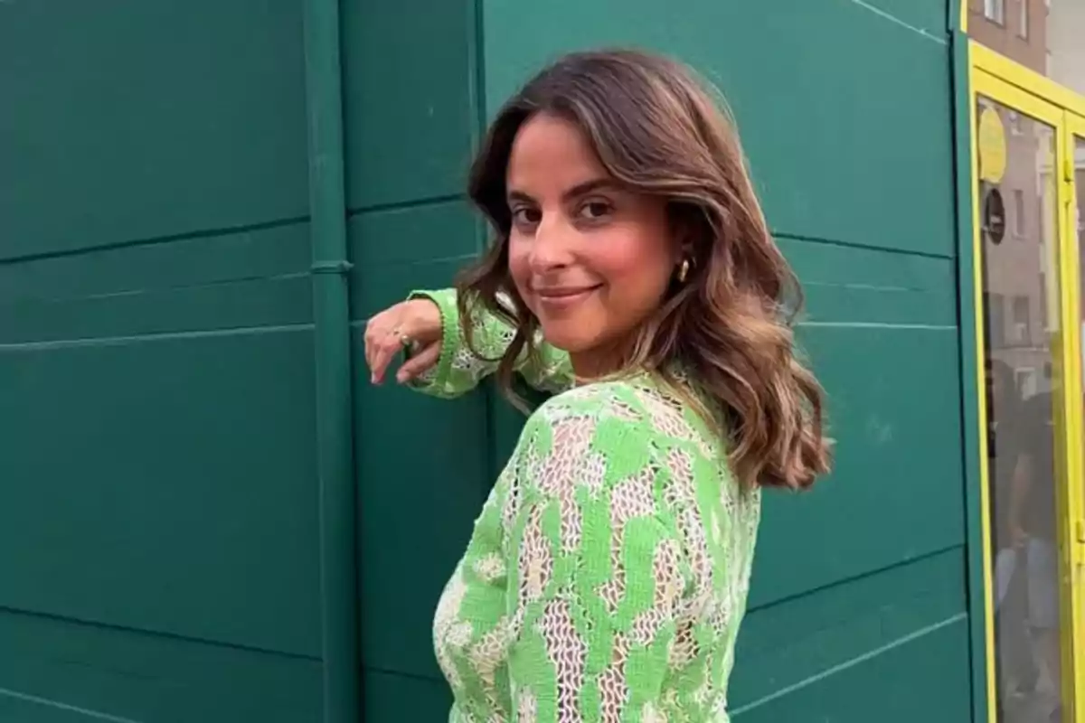 Mujer de cabello castaño y ondulado con suéter verde claro posando frente a una pared verde