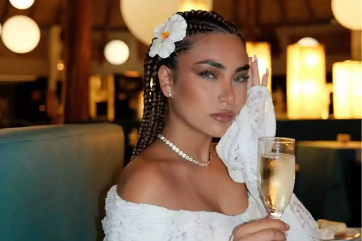 Mujer con trenzas y flor blanca en el cabello sosteniendo una copa de vino espumoso en un ambiente elegante iluminado Mujer con trenzas y flor blanca en el cabello sosteniendo una copa de vino espumoso en un ambiente elegante iluminado