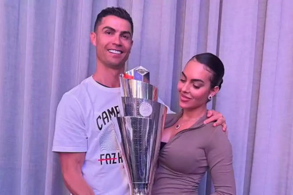 Pareja posando sonriente con un trofeo grande frente a una cortina gris